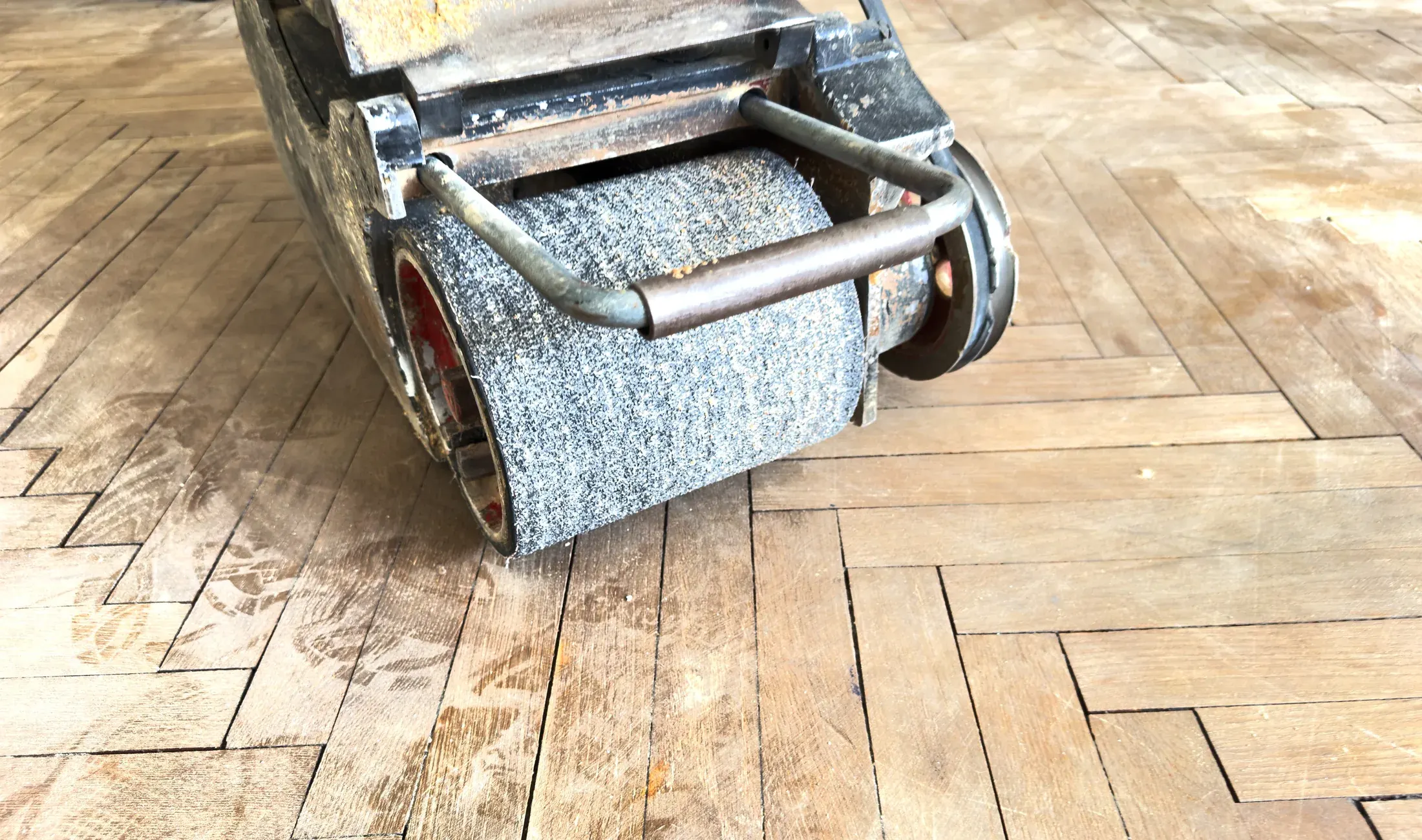 Drum floor sander refinishing a herringbone-pattern hardwood floor, no people visible